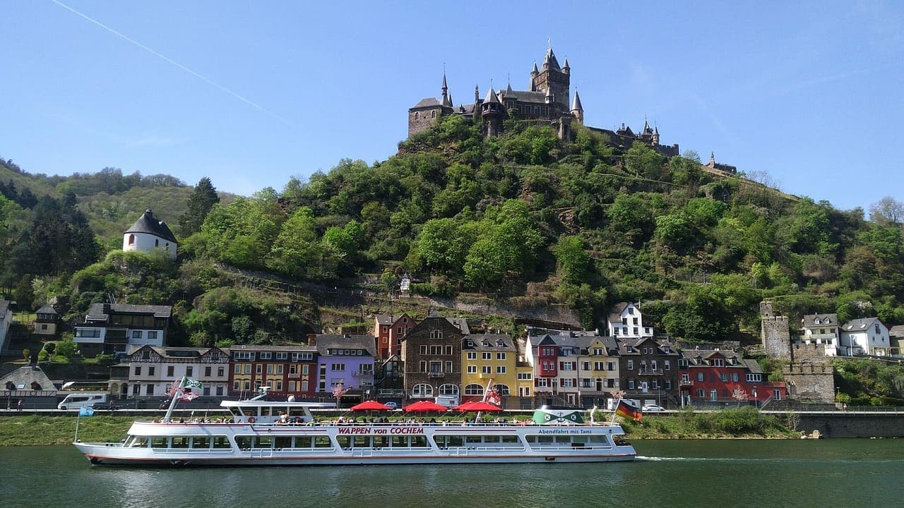 A river cruise boat sailing past a hilltop castle and traditional houses along the Rhine River in summer.