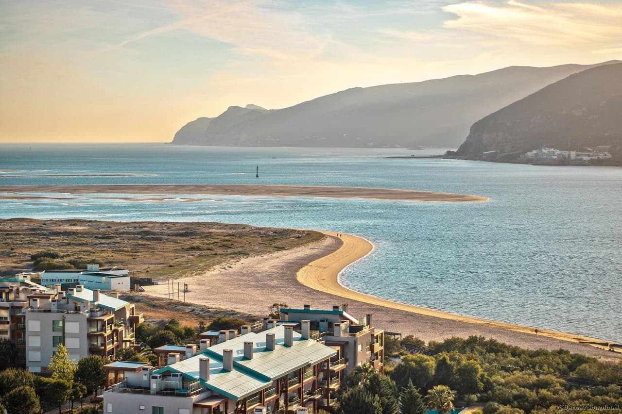 A wide coastal view in Portugal with a curved sandy beach, calm blue water and hills in the background at sunset.