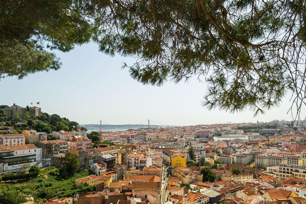 A panoramic view over Lisbon with red rooftops, historic buildings and the river in the distance under a clear sky.