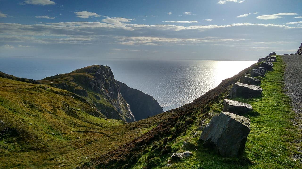 Coastal cliff path overlooking the Atlantic Ocean in Ireland, with grassy slopes and stone barriers leading along the edge.
