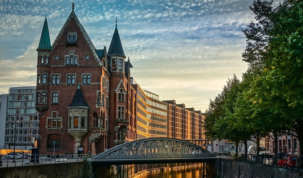 A historic red brick building beside a canal in a German city at sunset, with a small bridge and modern buildings in the background.
