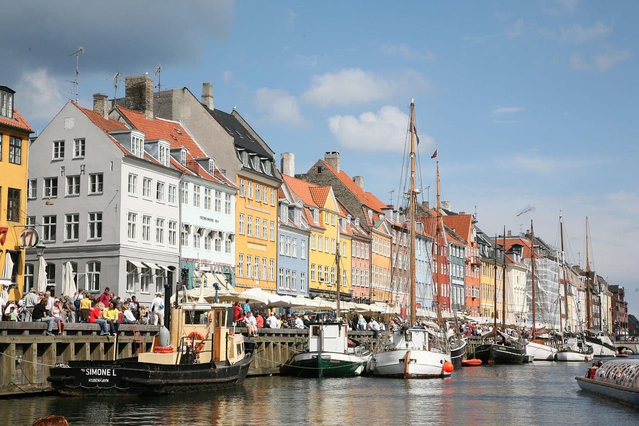 Historic harbour in Copenhagen lined with bright buildings and boats, with people enjoying the waterfront on a sunny day.