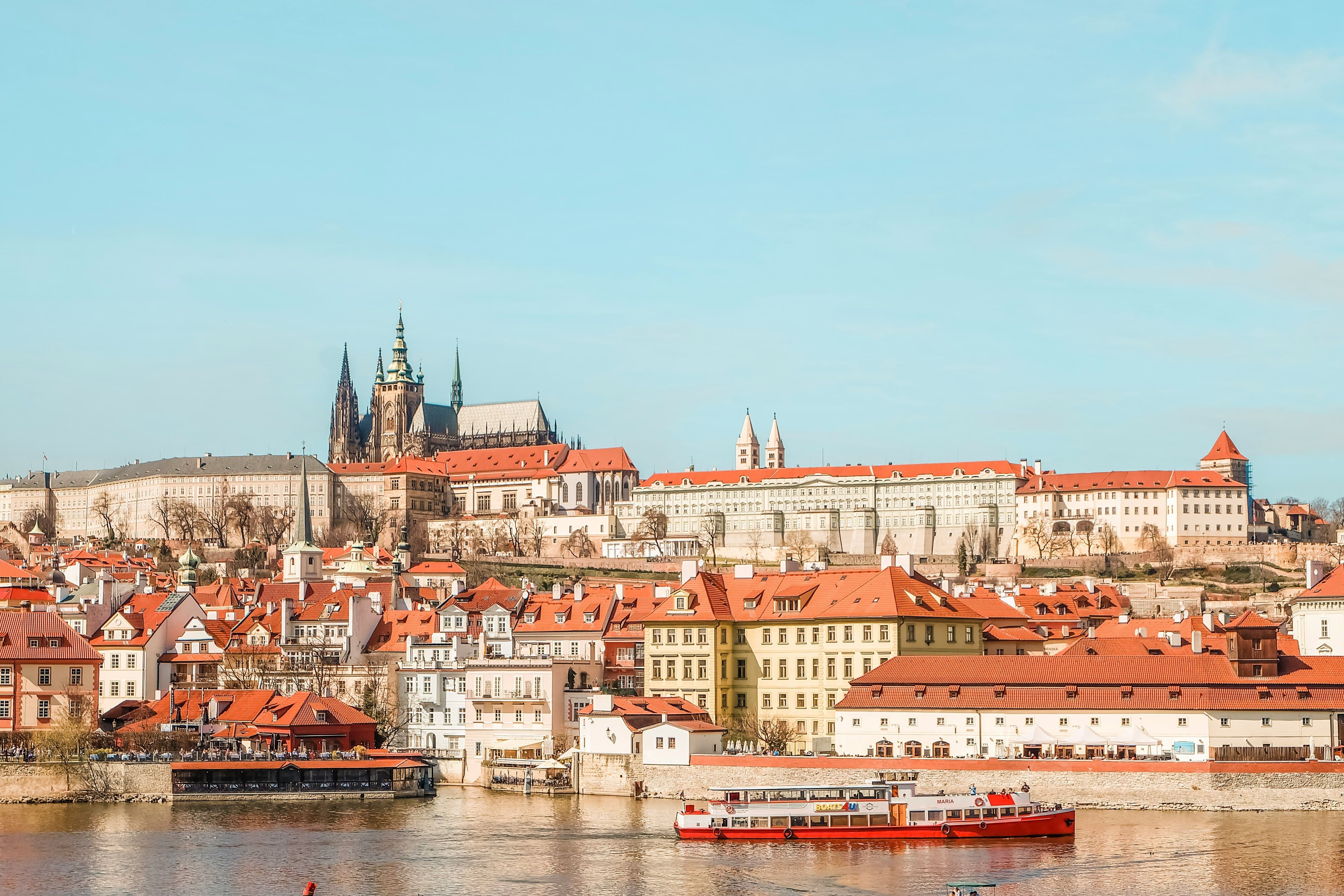 Picture of the city of Prague with a river and houses with orange roofs and the castle in the background on a clear day