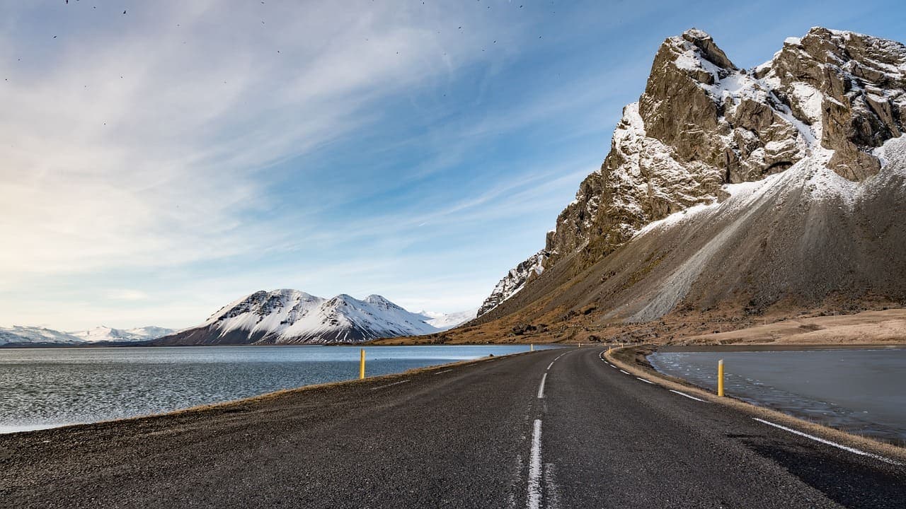 A winding Icelandic road running along a fjord with steep rocky mountains and snow patches under a blue sky.