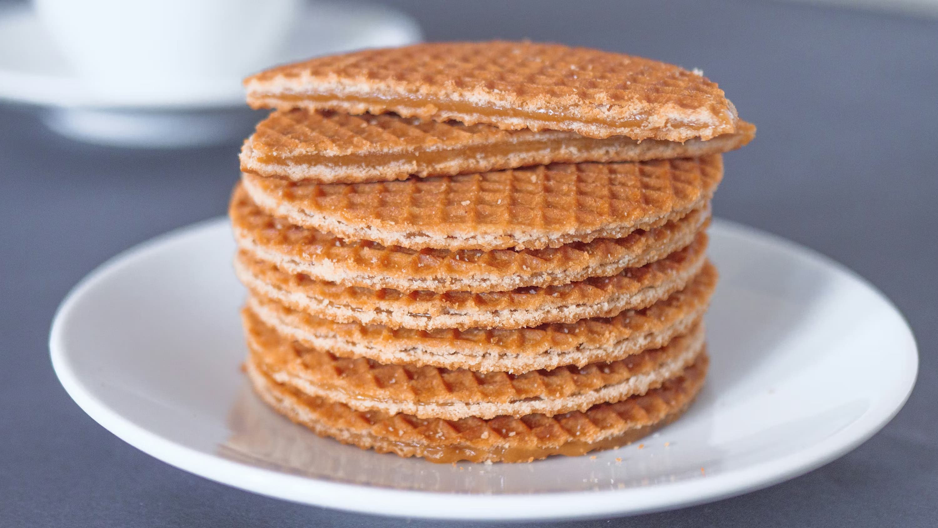 Stack of round, golden-brown stroopwafels on a white plate, with one partially broken on top. Background is softly blurred, creating a cozy, inviting feel.