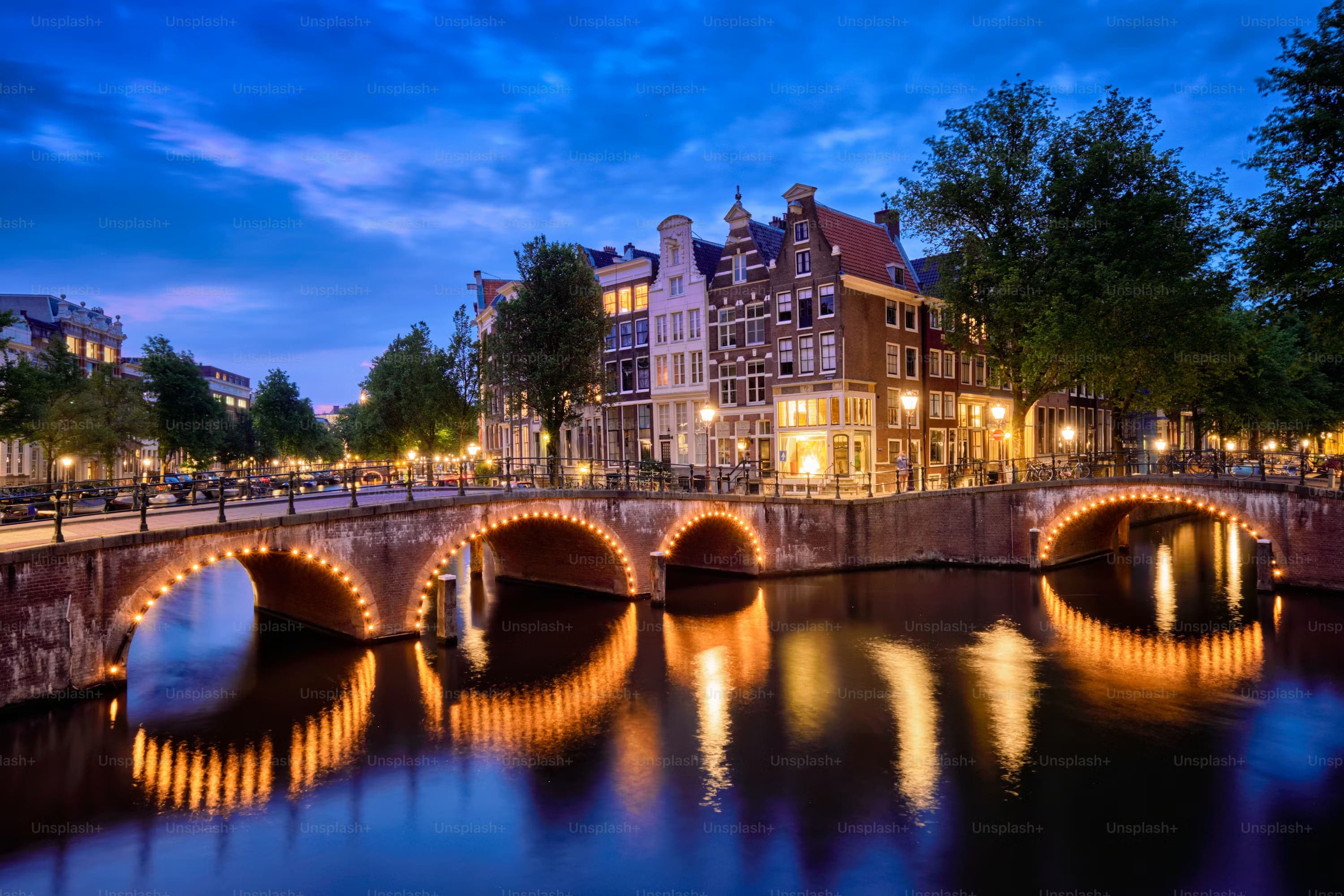 Illuminated bridges over an Amsterdam canal at dusk, with historic buildings and trees reflecting on the water, creating a serene ambiance.