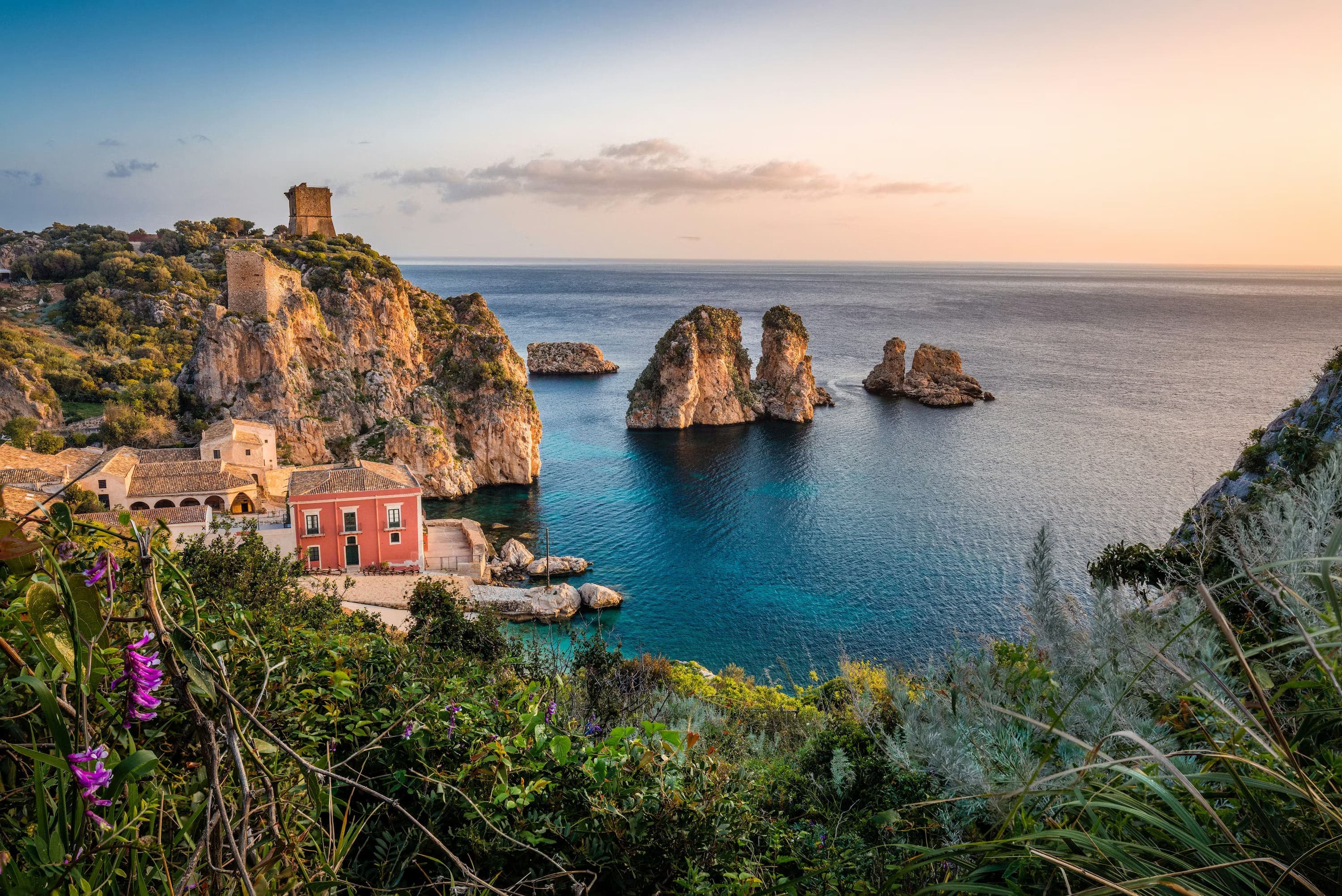 Sunset view of a coastal village with rocky cliffs and sea stacks, a historic watchtower, and pink flowers in the foreground, exuding tranquility.