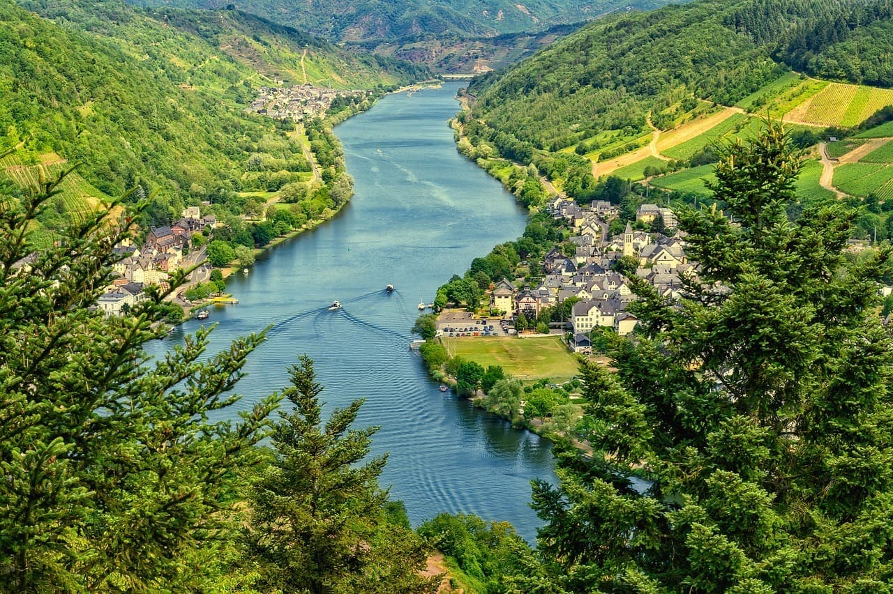 A wide river valley in Germany with green hills, vineyards and small villages along the water under a clear sky.