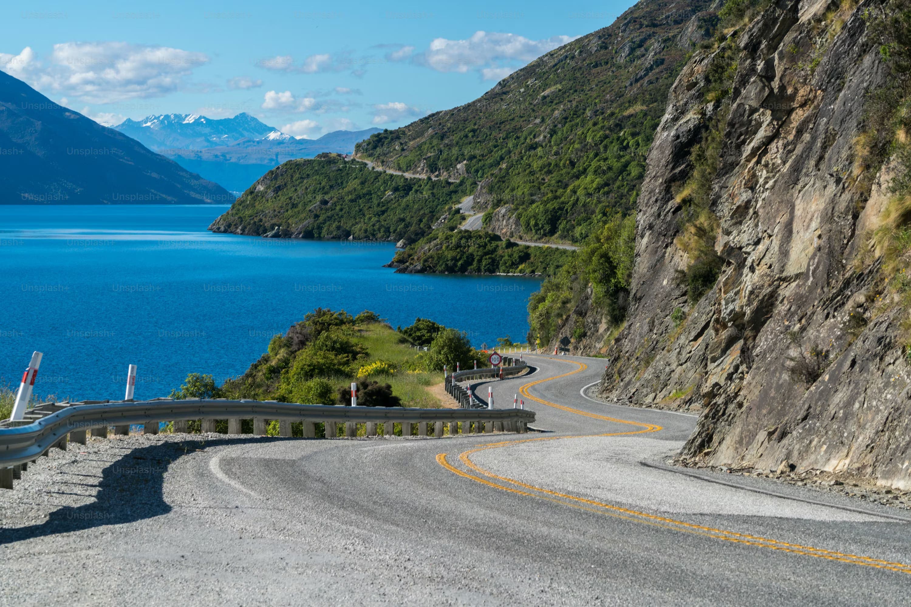 A winding mountain road curves alongside a bright blue lake, bordered by lush green hills under a clear sky with distant snowy peaks. Serene and scenic.