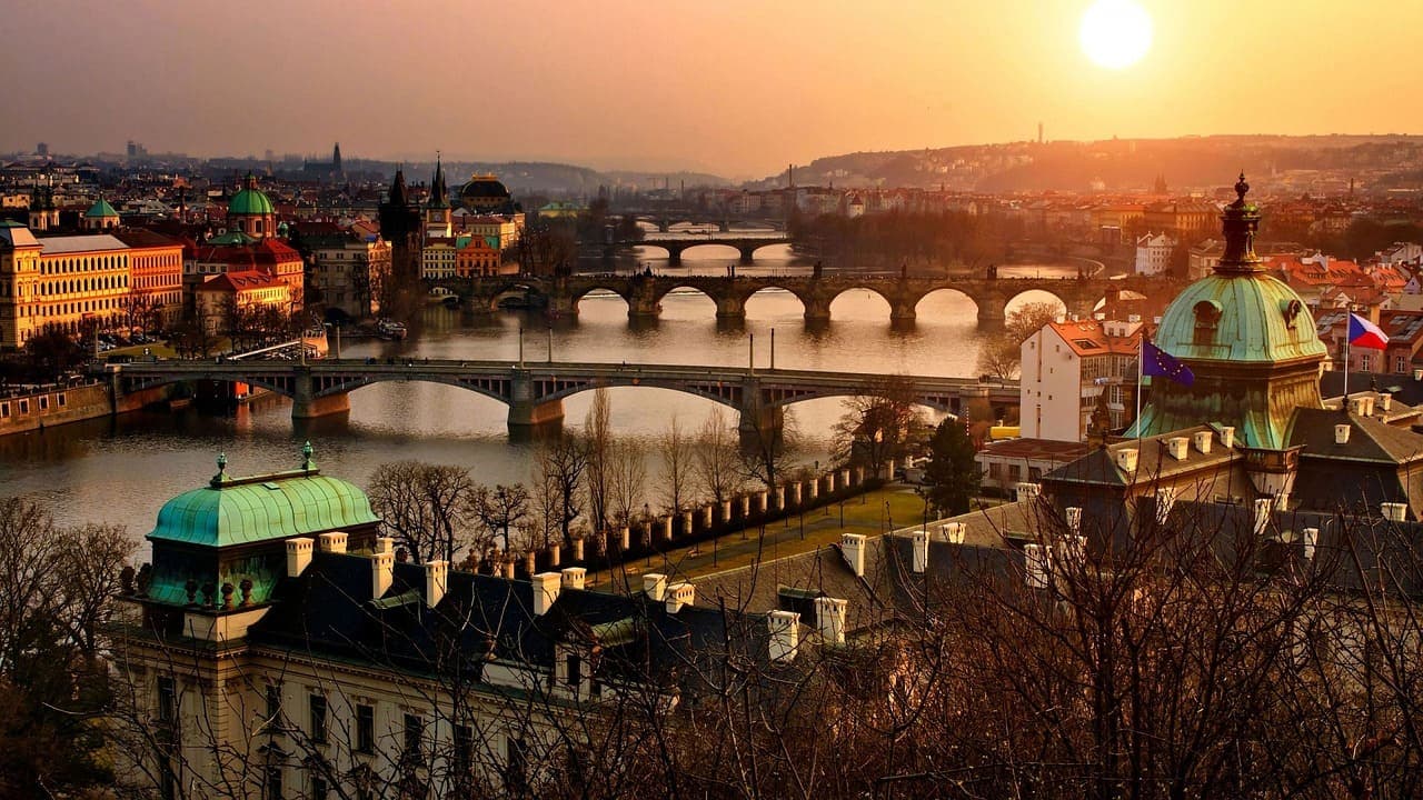 Sunset view of the Charles Bridge in Prague with historic buildings and the Vltava River reflecting the warm evening light.
