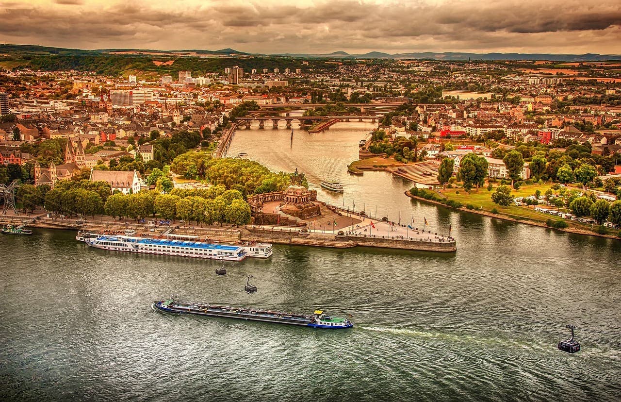 An aerial view of the Rhine River with boats sailing past a riverside city and bridges crossing the water.
