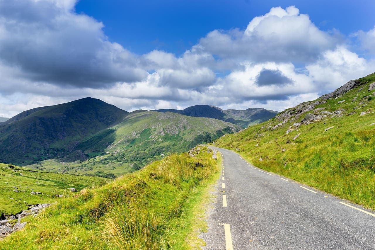 Winding rural road through green Irish countryside with hills and mountains in the distance beneath a blue sky with white clouds.