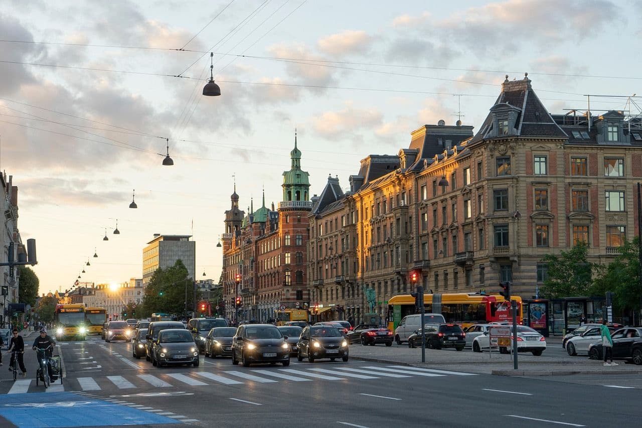 A wide city street in Copenhagen at sunset with historic buildings, cars and cyclists moving through the city, creating a lively urban atmosphere.