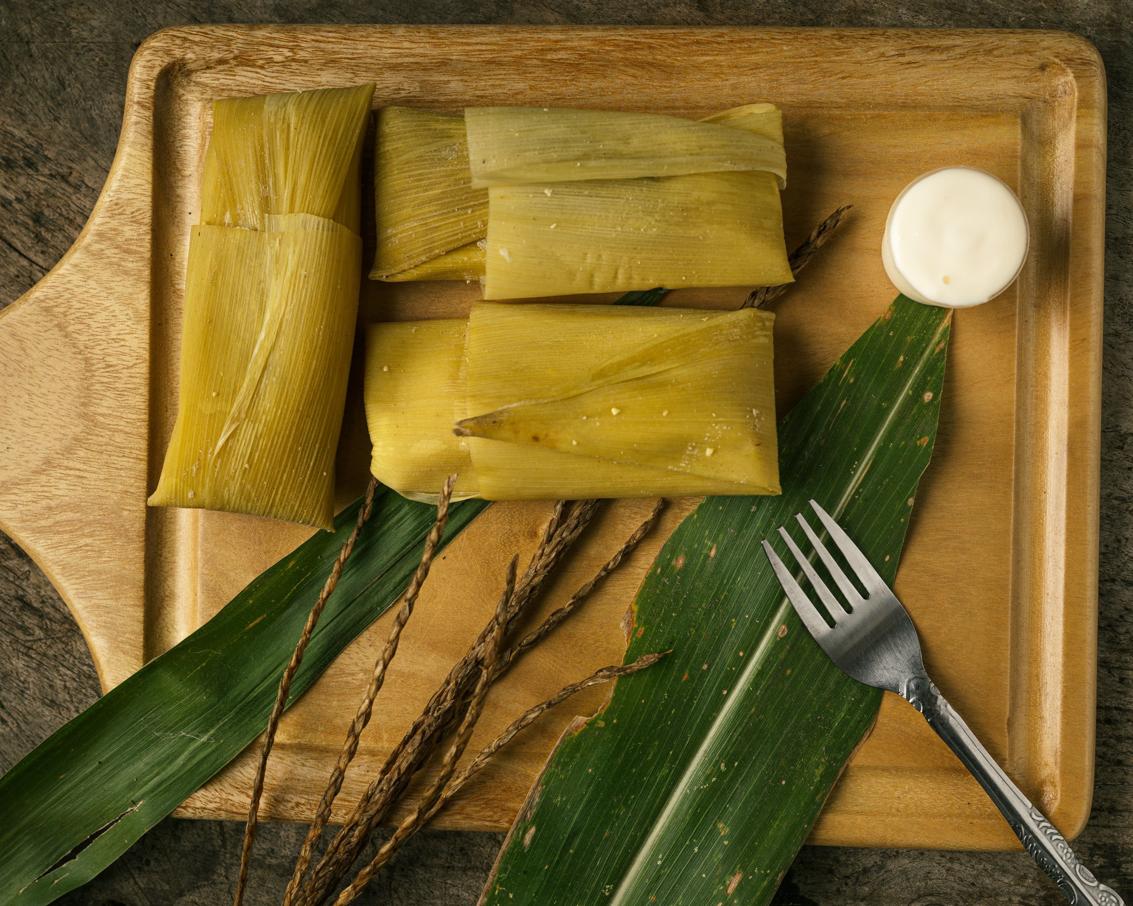 Traditional Guatemalan tamales wrapped in banana leaves served on a wooden tray with a fork and sauce.