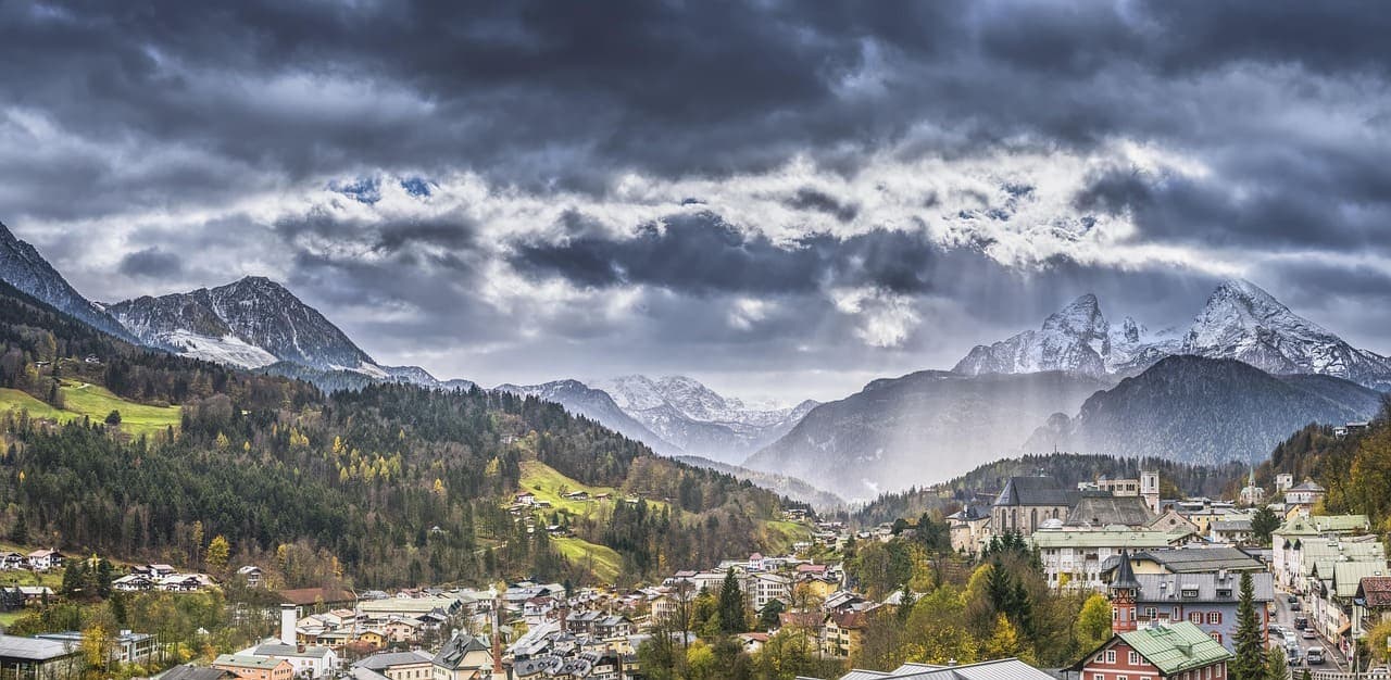 A panoramic view of a German town with mountains in the background, showing rooftops and alpine scenery under cloudy skies.
