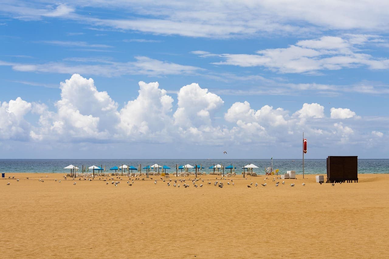 A golden sandy beach in Portugal with umbrellas, blue sky and soft white clouds above the Atlantic Ocean.