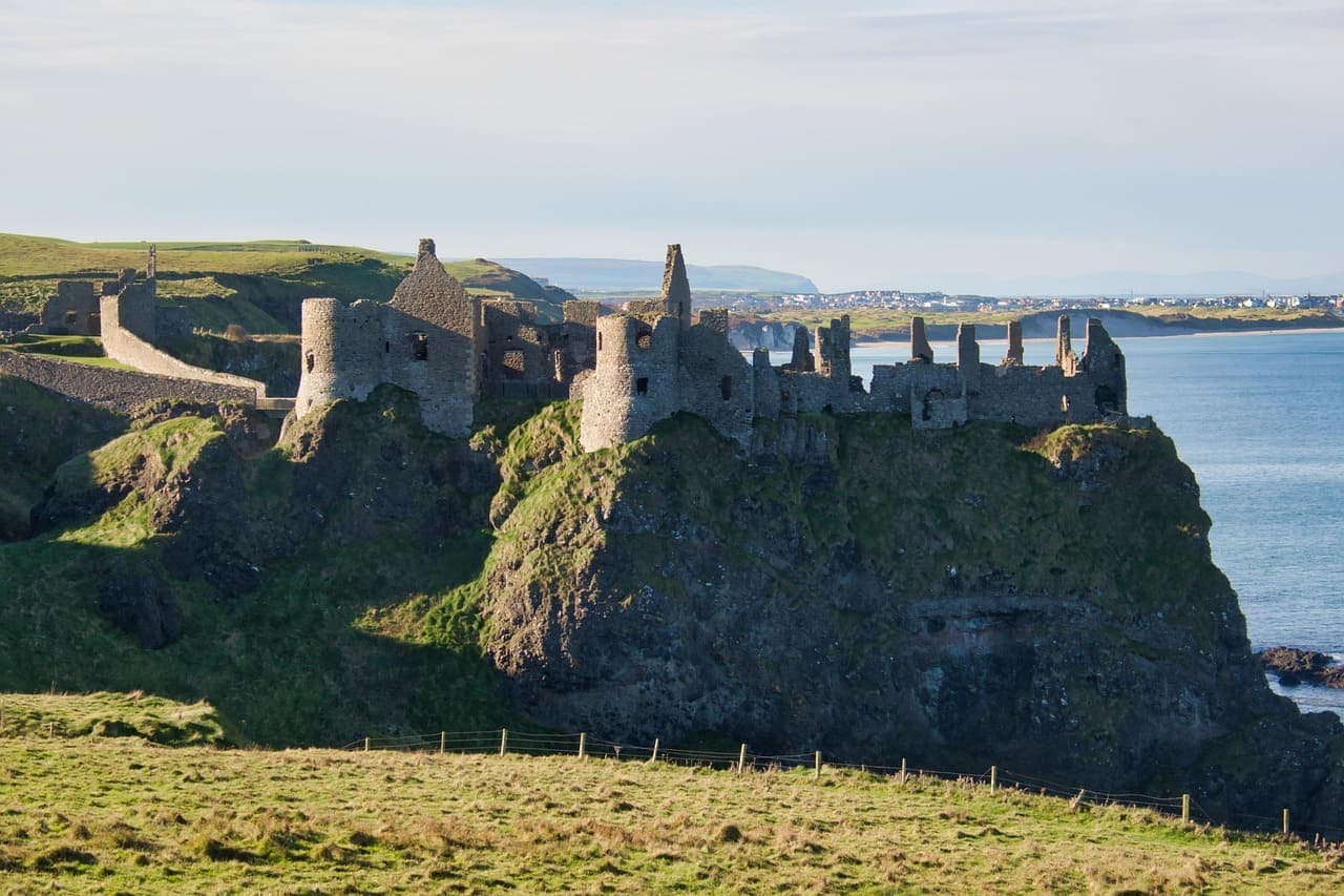 Medieval cliff top castle ruins overlooking the sea along the Northern Ireland coastline, surrounded by green grass and blue water.