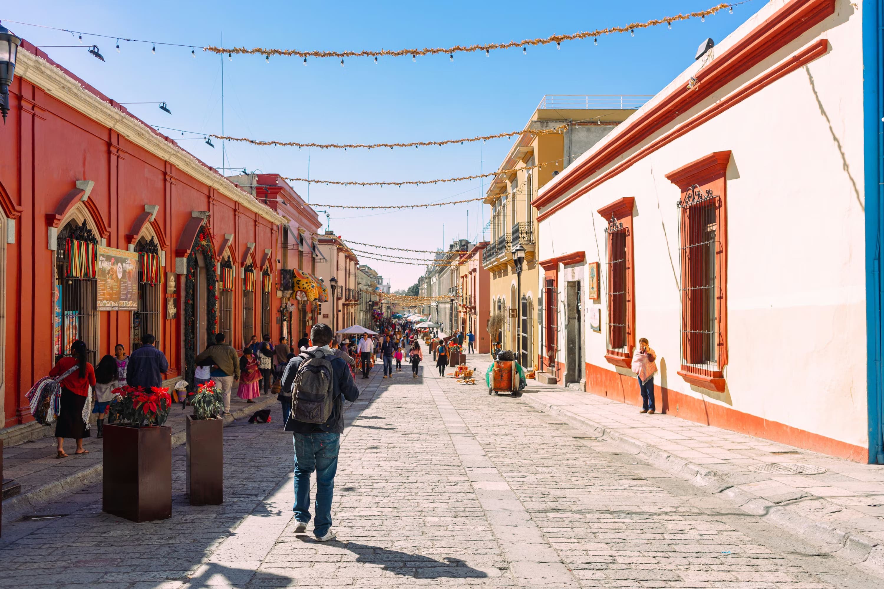 A vibrant street scene in Oaxaca, Mexico shows people walking past colorful colonial buildings; festive lights hang above, creating a lively, cheerful atmosphere.